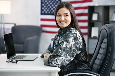 A female dressed in military fatigues crosses her arms and smiles at the camera while sitting at her work desk.