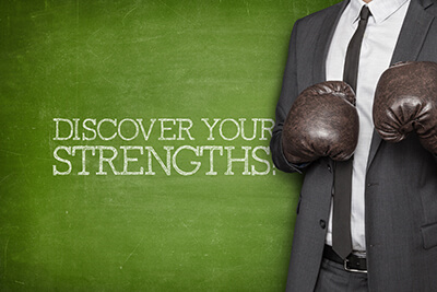 A person wearing business attire and boxing gloves stands next to the words “discover your strengths” stenciled in white chalk on a green chalkboard.