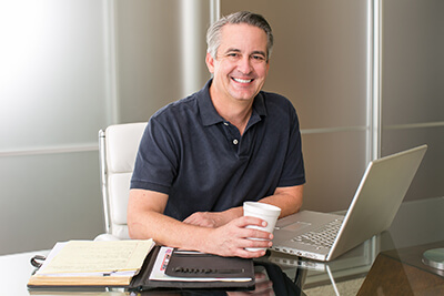 An entrepreneur smiles at the camera and holds a cup of coffee at their workspace.