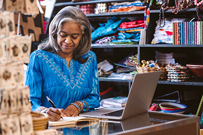 A smiling fabric-shop owner writing in a notebook while working from their laptop